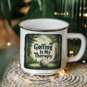May include: White enamel mug with a black rim and handle. The mug features a green and white graphic of a golf ball and trees with the text "Golfing Is My Therapy". The mug is on a woven surface.