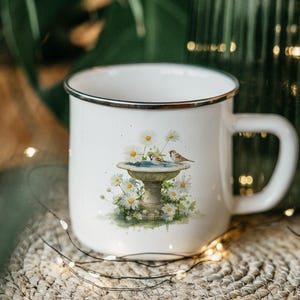 May include: White enamel mug with a black rim and handle. The mug features a watercolor illustration of a bird bath with daisies and two small birds. The mug is sitting on a woven surface.