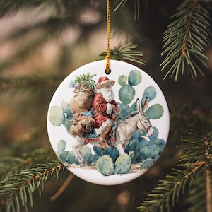 May include: Round Christmas ornament featuring Santa Claus riding a donkey through a desert landscape with cacti. Santa wears a red suit and hat, carrying a sack. The ornament is white with a gold string.