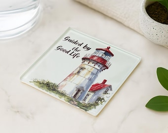 May include: Square glass coaster featuring a watercolor illustration of a lighthouse with a red roof and the text "Guided by the Good Life." The coaster is white with a clear border and is suitable for use with drinks.