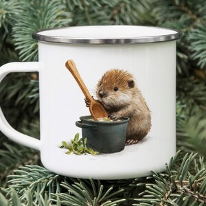 May include: White enamel mug with a silver rim featuring a watercolor illustration of a brown rodent stirring a pot with a wooden spoon. The mug has a white handle and is set against a backdrop of green pine needles.