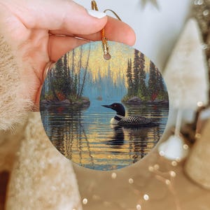 May include: Round ceramic ornament featuring a detailed painting of a loon swimming on a lake. The artwork depicts a serene landscape with trees and a colorful sky. The ornament has a gold string for hanging.