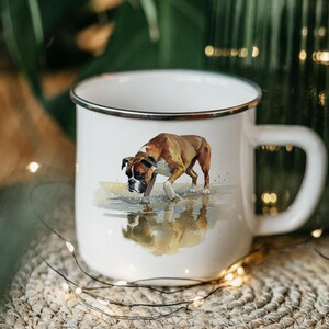 May include: White enamel mug with a black rim and handle. The mug features a watercolor painting of a Boxer dog walking in shallow water. The dog is brown and white, with a black muzzle. The mug is sitting on a woven surface.