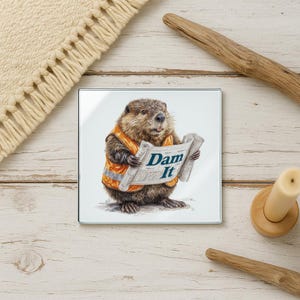 May include: Square glass coaster featuring a cartoon beaver wearing an orange safety vest and holding a blueprint. The blueprint reads "Dam It" in blue text. The coaster has a white background.