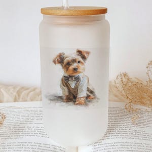 May include: A frosted glass tumbler with a wooden lid and straw. The tumbler features a watercolor illustration of a Yorkshire Terrier wearing a suit and bow tie. The tumbler is sitting on top of an open book.