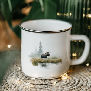 May include: White enamel mug with a black rim, featuring a serene landscape with a moose standing near a body of water. The design includes trees and a misty background, evoking a natural theme.