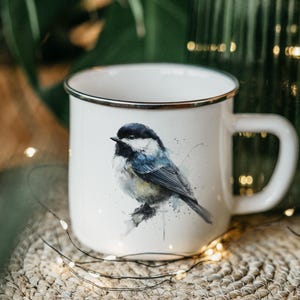 May include: White enamel mug with a black rim and handle. The mug features a watercolor painting of a small bird with black, blue, and yellow feathers. The mug is sitting on a woven mat.