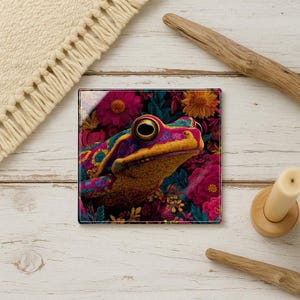 May include: Square glass coaster featuring a colorful frog design. The frog has yellow, blue, and pink markings, set against a backdrop of vibrant pink and orange flowers. A decorative item for home decor.