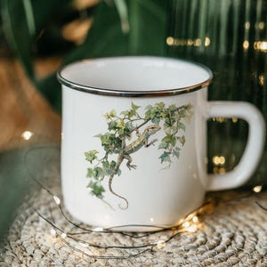 May include: White enamel mug with a silver rim and handle. The mug features a detailed illustration of a lizard perched on a leafy vine. The lizard is in shades of brown and green, and the leaves are green.