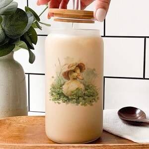 May include: A clear glass tumbler with a bamboo lid and straw, filled with a light brown beverage. The tumbler features a vintage-style illustration of a duck wearing a hat. The tumbler sits on a wooden tray.