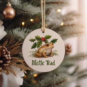 May include: Round ceramic Christmas ornament featuring a detailed illustration of a toad with holly and berries. The ornament has the text "Mistle Toad" in green. The ornament hangs from a gold string and is displayed on a Christmas tree.