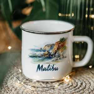 May include: White enamel mug with a silver rim, featuring a coastal watercolor painting and the word "Malibu" in blue script. The mug has a curved handle and is set on a woven surface.