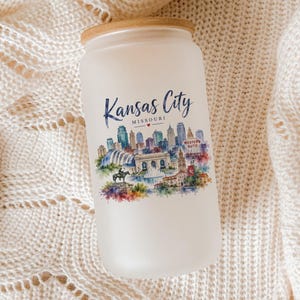 A white candle jar with a printed image of the Kansas City skyline on it, placed on a white knitted blanket.