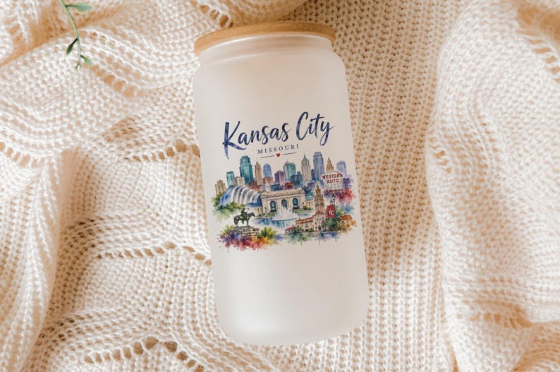 A white candle jar with a printed image of the Kansas City skyline on it, placed on a white knitted blanket.
