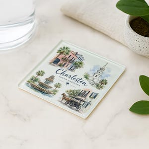 May include: Square glass coaster featuring watercolor illustrations of Charleston, South Carolina landmarks. The coaster includes images of buildings, a fountain, and a horse-drawn carriage, with the city's name and state printed on it.