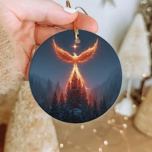 May include: Round Christmas ornament featuring a fiery phoenix rising above a forest. The phoenix is depicted in shades of orange and yellow against a dark blue background. The ornament has a gold string for hanging.
