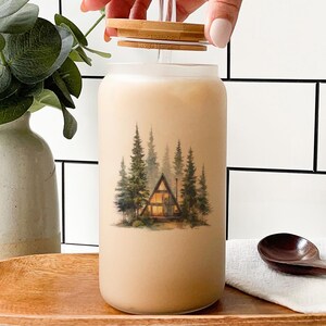 May include: A glass tumbler with a bamboo lid and straw, filled with a light-colored beverage. The tumbler features a watercolor illustration of a cabin in a forest. The scene includes green trees and a warm-lit cabin.