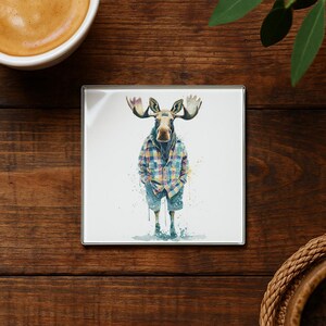 May include: Square coaster featuring a watercolor illustration of a moose wearing a plaid shirt and shorts. The moose has large antlers and is standing on a white background. The coaster has a silver border.