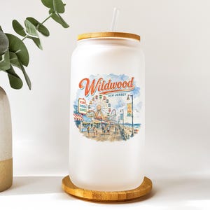 A glass jar with a printed design of a beach scene and a ferris wheel, placed on a wooden coaster.