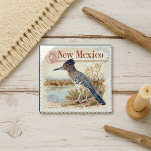 May include: Square coaster featuring a watercolor illustration of a roadrunner bird with the text "New Mexico". The coaster has a vintage postcard design with a light beige background and a decorative border.