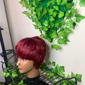 Op de afbeelding: Bordeauxkleurige pruik op een mannequinhoofd met een neusring. De mannequin staat op een zwart metalen rek, omringd door kunstgroen klimop. Een groene achtergrond is zichtbaar.