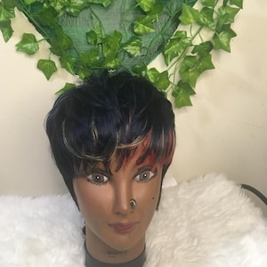 May include: A mannequin head with a short, layered wig. The wig features black hair with blue, red, and blonde highlights. The mannequin has a nose ring and is set against a white, fluffy background. Green ivy decorates the top.