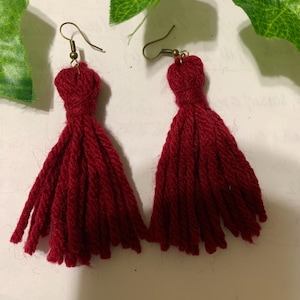 May include: A pair of handmade red tassel earrings. Each earring has a cluster of red yarn tassels hanging from a gold-coloured fish hook ear wire. The earrings are set against a neutral background with green leaf accents.