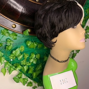 Può includere: Parrucca corta, nera e bianca, a strati, esposta su una testa di manichino. La parrucca ha una scriminatura laterale con una sezione bianca. Il manichino è su un supporto verde e indossa una collana nera.