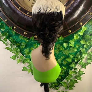 Può includere: Parrucca bicolore nera e bianca, acconciata con un taglio corto e a strati sulla parte superiore e ciocche ricci più lunghe sul retro. La parrucca è esposta su una testa di manichino con un busto verde.