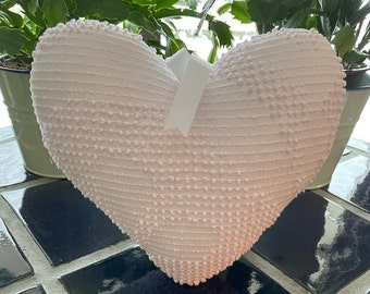 May include: A light pink heart-shaped decorative pillow with a textured surface. A white ribbon is attached to the top of the pillow. The pillow is sitting on a dark blue and black tiled surface.