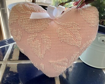 May include: A pink heart-shaped pillow with a white satin ribbon tied around it. The pillow has a textured surface and is sitting on a blue and white tiled surface.
