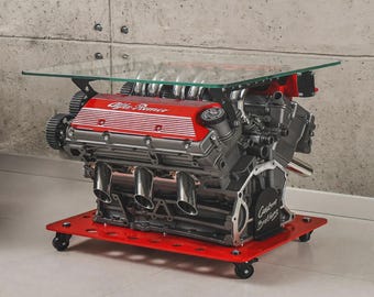 May include: A coffee table featuring a glass top supported by a repurposed Alfa Romeo engine block. The engine is painted red and grey, mounted on a red metal base with four black caster wheels. The text Custom Brothers is visible.