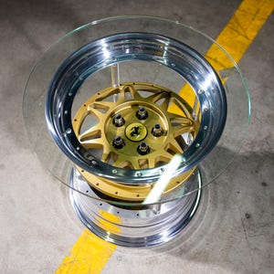 May include: A circular glass-topped table featuring a base constructed from a metallic car wheel rim. The rim has a polished chrome outer edge and a gold-colored multi-spoke center section with five lug nuts and a central emblem.