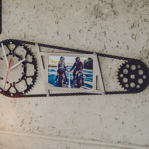 Może przedstawiać: Czarny metalowy zegar z łańcuchem i zębatkami, z fotografią pary jadącej na motocyklach. Zegar jest zamontowany na jasnoszarej ścianie.