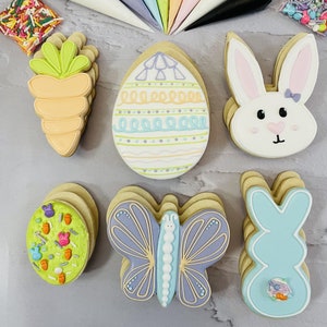 May include: A set of Easter-themed sugar cookies decorated with colorful icing. The cookies include a butterfly, a carrot, an egg, and two bunnies. The cookies are arranged on a light gray marble surface with piping bags and sprinkles in the background.