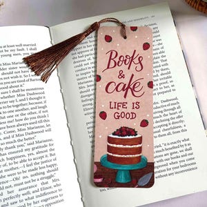 May include: A bookmark with the words "Books & cake" and "Life is Good". The bookmark has illustrations of strawberries and a layered cake on a teal cake stand. A brown tassel hangs from the top. The bookmark is resting on an open book.