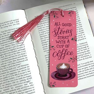 May include: A pink bookmark featuring the text "All good stories start with a cup of coffee" and a coffee cup illustration. It has a pink tassel and is in an open book.