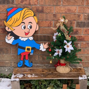 May include: A cartoon elf figurine with a blue hat, blue shirt, red pants, and blue boots stands on a wooden bench next to a small artificial Christmas tree decorated with red berries, white flowers, and gold bells. The tree is in a burlap sack.