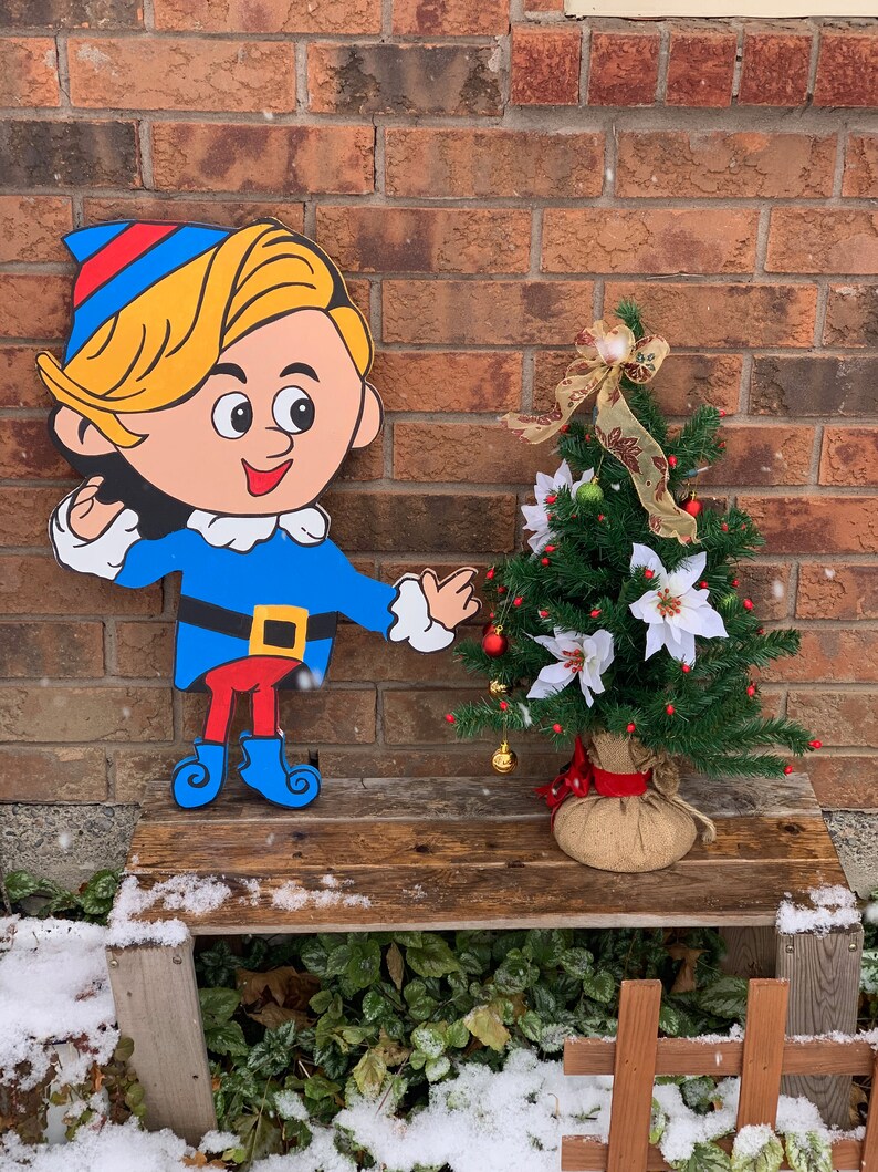 May include: A cartoon elf figurine with a blue hat, blue shirt, red pants, and blue boots stands on a wooden bench next to a small artificial Christmas tree decorated with red berries, white flowers, and gold bells. The tree is in a burlap sack.