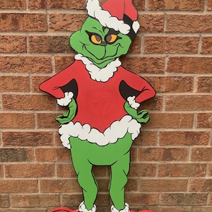 Peut inclure: Un personnage vert du Grinch portant un costume de Père Noël rouge avec des bordures blanches et un chapeau de Père Noël. Le Grinch est debout, les bras croisés, et a une expression de mécontentement sur son visage.