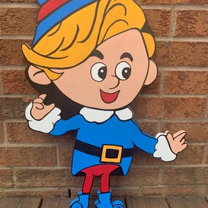 May include: A wooden cutout of a cartoon elf with blonde hair, wearing a blue and red hat, a blue shirt with a white collar, a black belt with a yellow buckle, red pants, and blue boots. The elf is smiling and has a friendly expression.