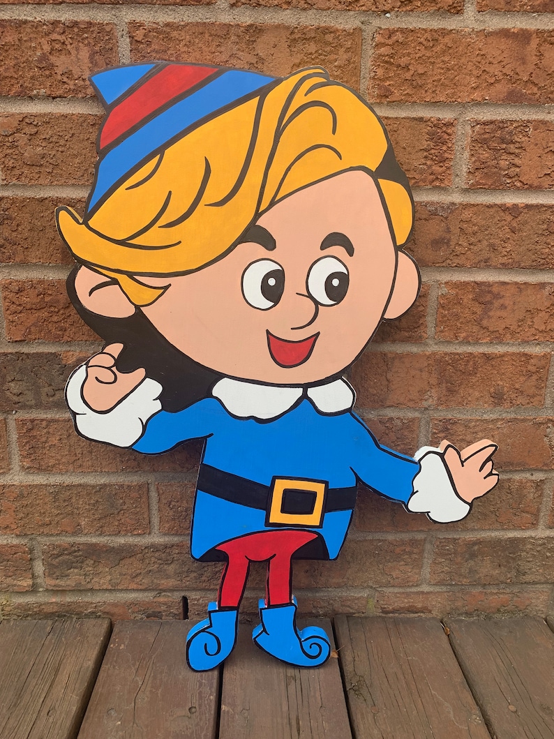 May include: A wooden cutout of a cartoon elf with blonde hair, wearing a blue and red hat, a blue shirt with a white collar, a black belt with a yellow buckle, red pants, and blue boots. The elf is smiling and has a friendly expression.