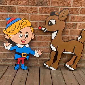 Op de afbeelding: Houten uitsparingen van een cartoon elf en een rendier. De elf draagt een blauwe outfit met een rode en blauwe hoed. Het rendier is bruin met witte accenten en een rode neus. De figuren leunen tegen een bakstenen muur.
