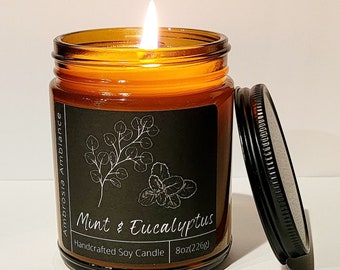 May include: A lit, amber-colored glass jar candle with a black label featuring botanical illustrations and the text "Mint & Eucalyptus." The candle is handcrafted soy and weighs 8oz (226g). The black lid is open beside the jar.