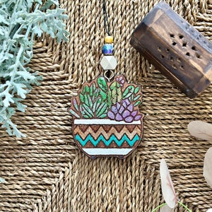May include: A wooden pendant necklace featuring a painted succulent plant in a pot with a brown, white, and turquoise chevron pattern. The pendant is attached to a black cord with a white bead and blue and yellow beads.