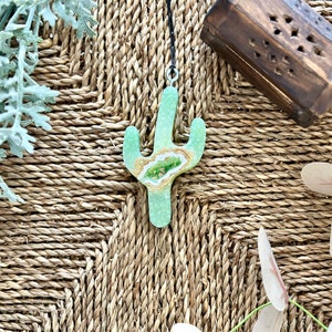 Könnte beinhalten: Ein grüner und goldener Kaktus-Anhänger aus Harz mit einem klaren Geoden-Design. Der Anhänger hängt an einer schwarzen Schnur und befindet sich auf einer gewebten Oberfläche.