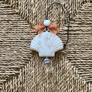 Könnte beinhalten: Ein weißer Muschelanhänger mit einer pfirsichfarbenen Schleife und einem Kristalltropfen, der an einem schwarzen Band hängt. Der Anhänger befindet sich auf einem geflochtenen Strohhintergrund.