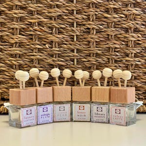 Op de afbeelding: Een verzameling glazen flessen met houten doppen en houten kogelverspreiders. Elke fles heeft een label met een geurnaam, zoals "Luxury Leather" en "Velvet Latte". De achtergrond is een gevlochten mand.