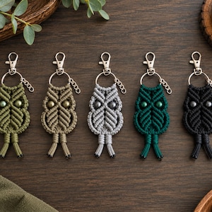 May include: Five macrame owl keychains arranged in a row, each featuring a silver-toned lobster clasp and ring. The owls are crafted from cord in olive green, tan, light gray, dark green, and black, with two round beads serving as eyes.