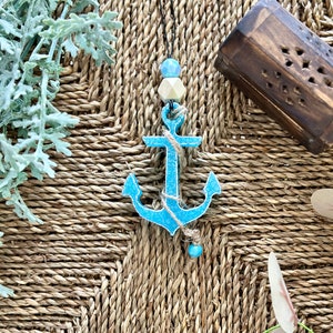 Op de afbeelding: Een blauwe houten anker met een touw en kralen die eraan hangen. De anker ligt op een geweven bruine achtergrond.